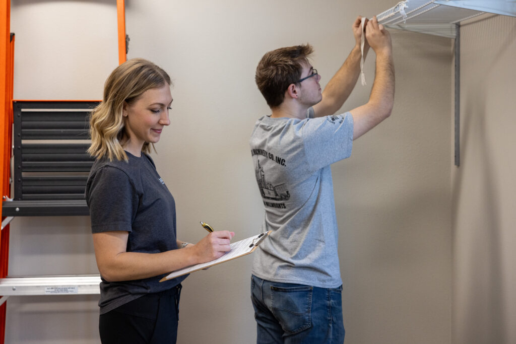 A woman takes notes while a man measures a wall with a tape measure.